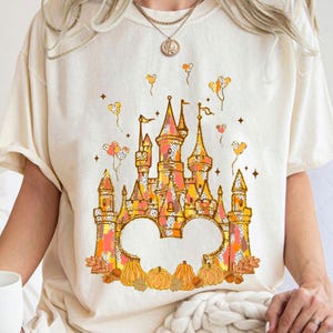 May include: Cream-colored t-shirt featuring a colorful castle design with orange, pink, and yellow hues. The castle is adorned with flags and surrounded by pumpkins, leaves, and balloons. A whimsical and festive graphic tee.