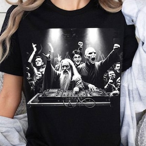 May include: Black t-shirt featuring a black and white graphic of a DJ set with a crowd. The DJ set includes a wizard, Harry Potter, and Voldemort. The crowd is cheering with their hands in the air.
