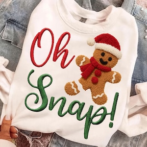 May include: White sweatshirt with the embroidered phrase "Oh Snap!" in red and green. A gingerbread man with a red scarf and Santa hat is featured on the front. The sweatshirt is a festive holiday garment.