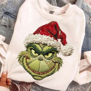 Puede incluir: Sudadera blanca con la cara verde del Grinch con un gorro de Papá Noel rojo con ribete blanco y un pompón. La cara del Grinch está detallada con varias texturas verdes y ojos amarillos, creando un diseño festivo navideño.