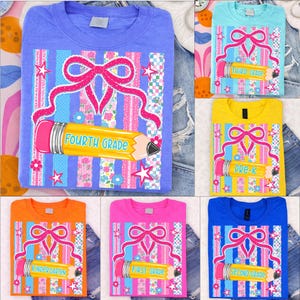 May include: Assorted-color t-shirts with school-themed designs. Each shirt features a large pencil graphic with the grade level printed on it, along with a pink bow and decorative stripes. The shirts are in various colors, including blue, orange, and yellow.