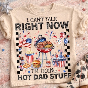 May include: Beige t-shirt with a graphic featuring a barbecue scene with an American flag, grill, food, and the text "I CAN'T TALK RIGHT NOW I'M DOING HOT DAD STUFF". The design includes fireworks, balloons, and a checkered border.