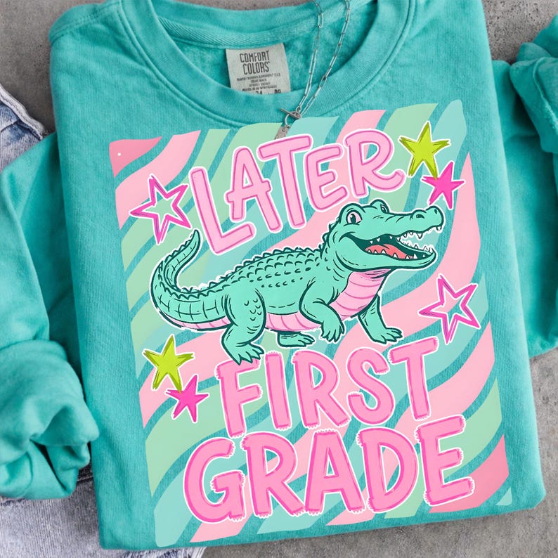 Peut inclure: Sweat-shirt turquoise avec un motif d'alligator de dessin anim&eacute; et les mots "LATER FIRST GRADE" en rose. Le motif comprend des rayures roses et vertes et des &eacute;toiles. Un collier argent&eacute; est visible.