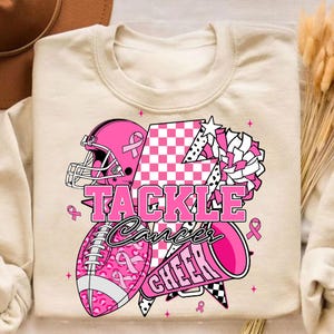 May include: Cream-coloured sweatshirt with a pink graphic design. The design includes a football helmet, football, and megaphone, with the words "TACKLE Cancer CHEER". Pink ribbons and stars are also featured in the design.