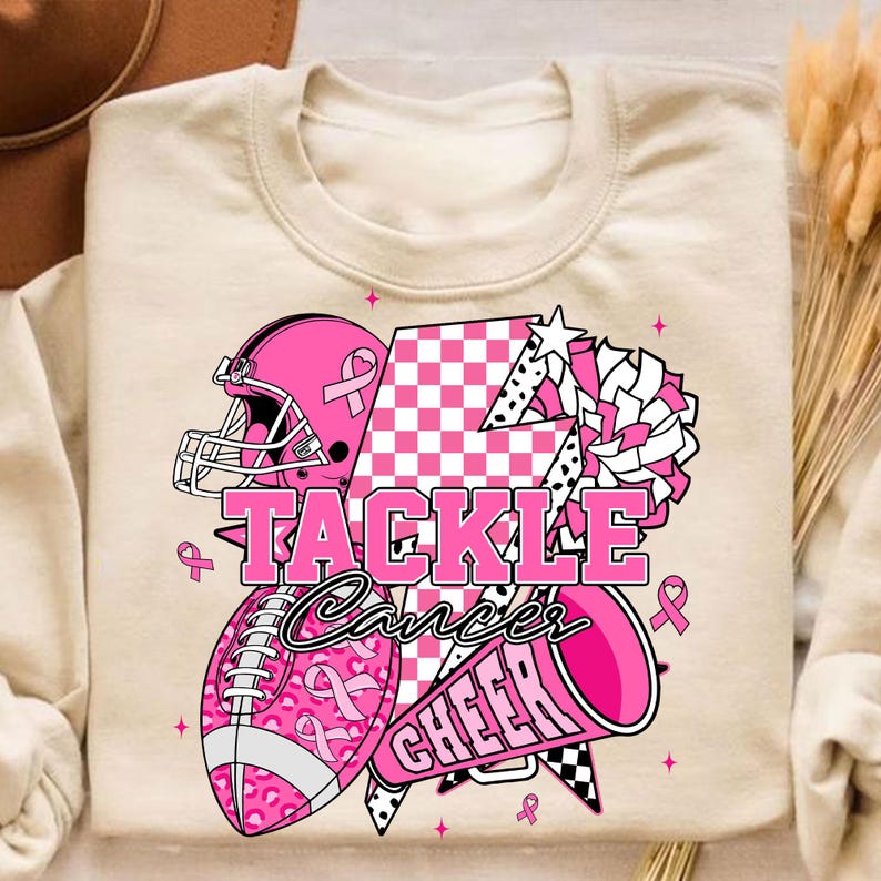 May include: Cream-coloured sweatshirt with a pink graphic design. The design includes a football helmet, football, and megaphone, with the words "TACKLE Cancer CHEER". Pink ribbons and stars are also featured in the design.