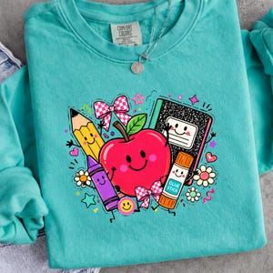 May include: A teal long-sleeve sweatshirt featuring a graphic print of a smiling red apple, a yellow pencil, a purple crayon, a black and white composition notebook, and a glue stick, all with faces, surrounded by pink bows and small stars.