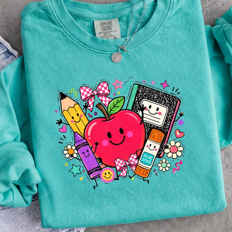 May include: A teal long-sleeve sweatshirt featuring a graphic print of a smiling red apple, a yellow pencil, a purple crayon, a black and white composition notebook, and a glue stick, all with faces, surrounded by pink bows and small stars.