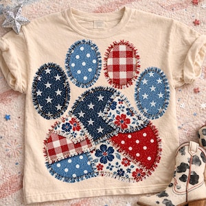 May include: Cream-colored t-shirt featuring a patriotic paw print design. The paw print is composed of patches in red, white, and blue, with star and polka dot patterns. A pair of white and black cowboy boots are visible.