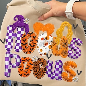 May include: Beige sweatshirt with the words "Hocus Pocus" in a patchwork design. The letters feature Halloween-themed patterns, including bats, spiderwebs, and checkered patterns. The design incorporates orange, purple, and white colours.