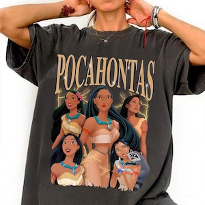 Puede incluir: Camiseta gris oscuro con la palabra "POCAHONTAS" en letras doradas sobre un gráfico del personaje. El gráfico incluye múltiples imágenes del personaje con un mapache. La camiseta está hecha de un material suave.