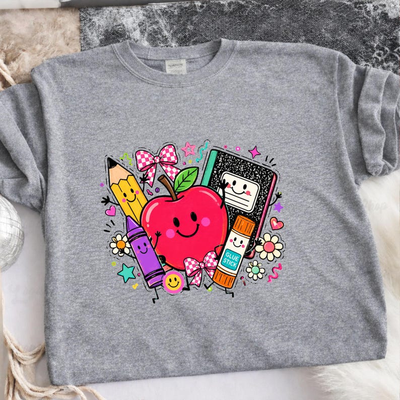 May include: A heather gray t-shirt featuring a colorful graphic of a smiling red apple surrounded by a yellow pencil, a purple crayon, a black and white composition notebook, and an orange glue stick, accented with pink bows and small flowers.