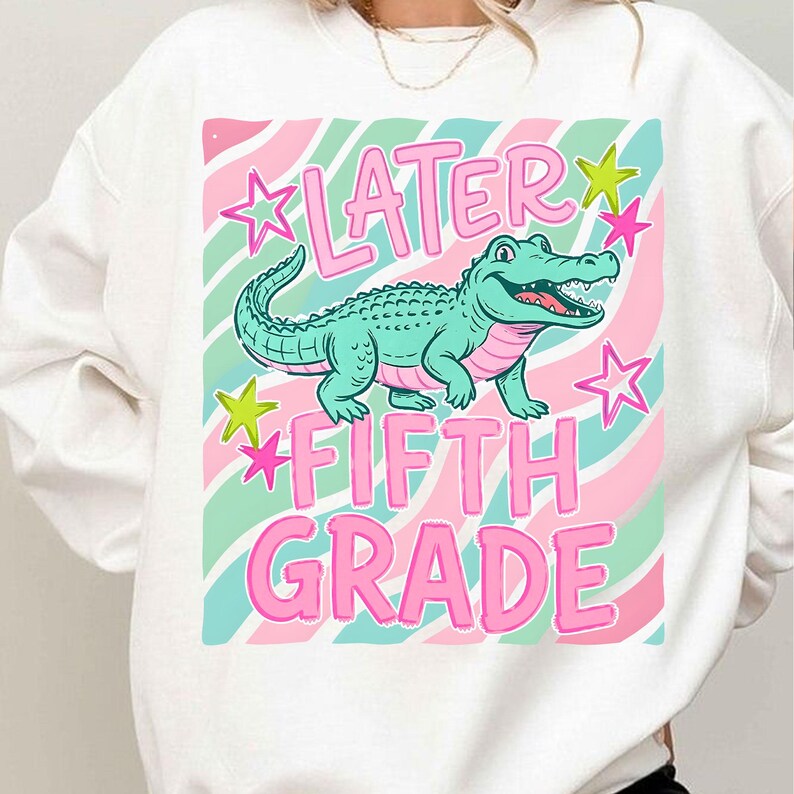 Peut inclure: Sweat-shirt blanc avec un motif graphique repr&eacute;sentant un alligator turquoise, des &eacute;toiles roses et le texte "LATER FIFTH GRADE" en rose. Le fond pr&eacute;sente un motif &agrave; rayures roses et turquoise.