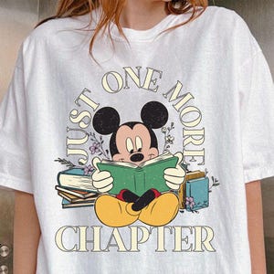 Op de afbeelding: Wit T-shirt met een cartoon afbeelding van Mickey Mouse die een boek leest. De tekst "Just One More Chapter" staat op het shirt gedrukt.