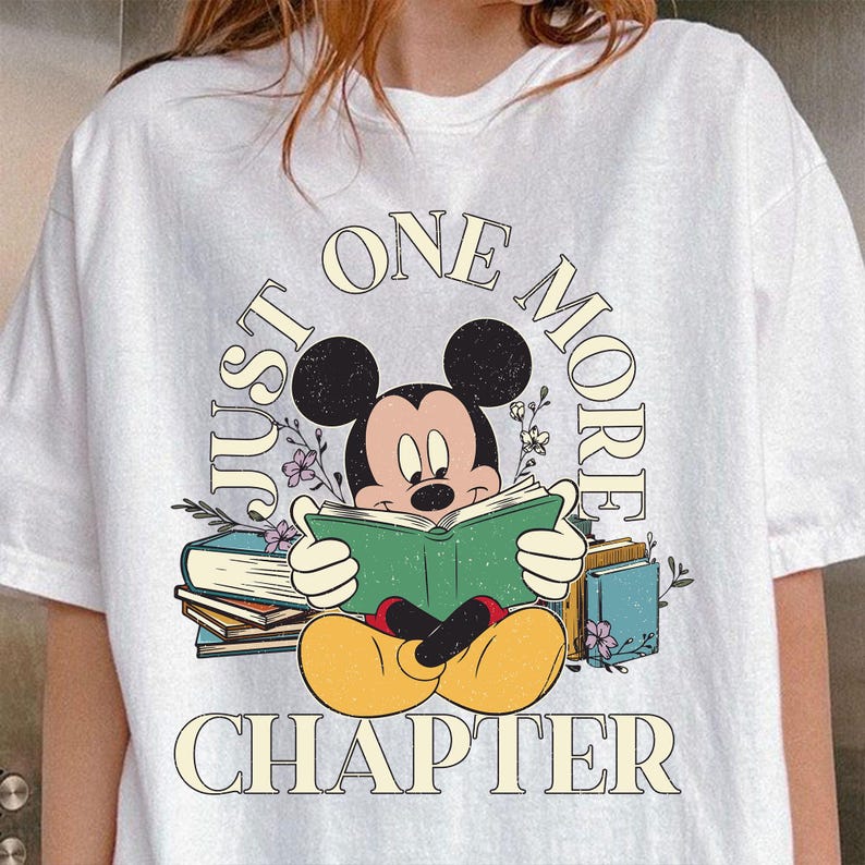 Op de afbeelding: Wit T-shirt met een cartoon afbeelding van Mickey Mouse die een boek leest. De tekst "Just One More Chapter" staat op het shirt gedrukt.