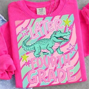 Peut inclure: Un sweat-shirt rose vif avec un motif d'alligator turquoise et l'inscription "LATER FOURTH GRADE". Le design comprend des rayures roses et turquoise et des &eacute;toiles. Un collier argent&eacute; est visible.