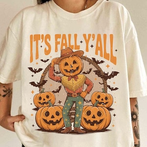 May include: Cream-colored t-shirt with a fall-themed graphic. The design features a pumpkin-headed cowboy with a rope lasso, surrounded by jack-o'-lanterns and bats. The text "IT'S FALL Y'ALL" is printed above the cowboy.