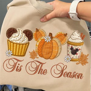 May include: Beige sweatshirt with an embroidered fall-themed design. The design includes a cupcake, pumpkins, and a coffee cup, all with a Mickey Mouse theme. The text "Tis The Season" is also embroidered in a brown script.