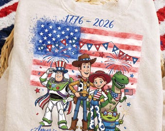 May include: A beige sweatshirt featuring a graphic of the American flag and characters from the Toy Story franchise. The design includes the text "1776-2026" and "America - 250th Anniversary". The design is in red, white, and blue.