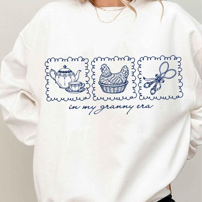 May include: White sweatshirt with a blue graphic featuring a teapot and teacup, a hen in a basket, and scissors. The text "in my granny era" is below the graphics. The design is in a rectangular frame.