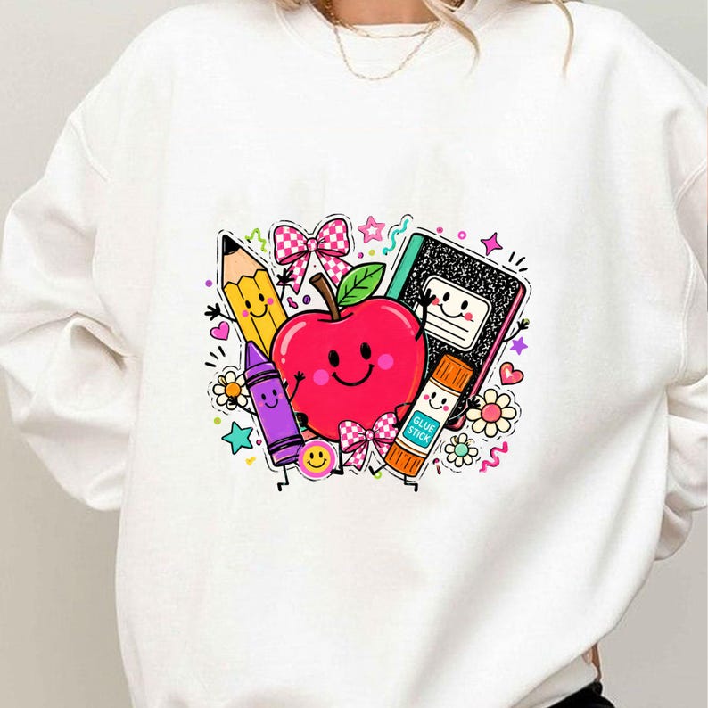 May include: A white crewneck sweatshirt featuring a colorful graphic print of a smiling red apple surrounded by a yellow pencil, a purple crayon, a black composition notebook, and an orange glue stick labeled GLUE STICK, accented with pink bows.