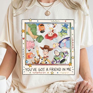 Puede incluir: Camiseta color crema con un dise&ntilde;o gr&aacute;fico colorido de personajes de Toy Story, incluyendo a Woody y Buzz Lightyear. El dise&ntilde;o est&aacute; enmarcado e incluye el texto "YOU'VE GOT A FRIEND IN ME".