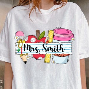 K&ouml;nnte beinhalten: Wei&szlig;es T-Shirt mit einem farbenfrohen Grafikdesign mit Elementen zum Thema Schule. Das Design umfasst einen roten Apfel, einen Bleistift, ein Lineal und einen rosa Cupcake. Der Text "Mrs. Smith" steht auf einem linierten Papierbanner.