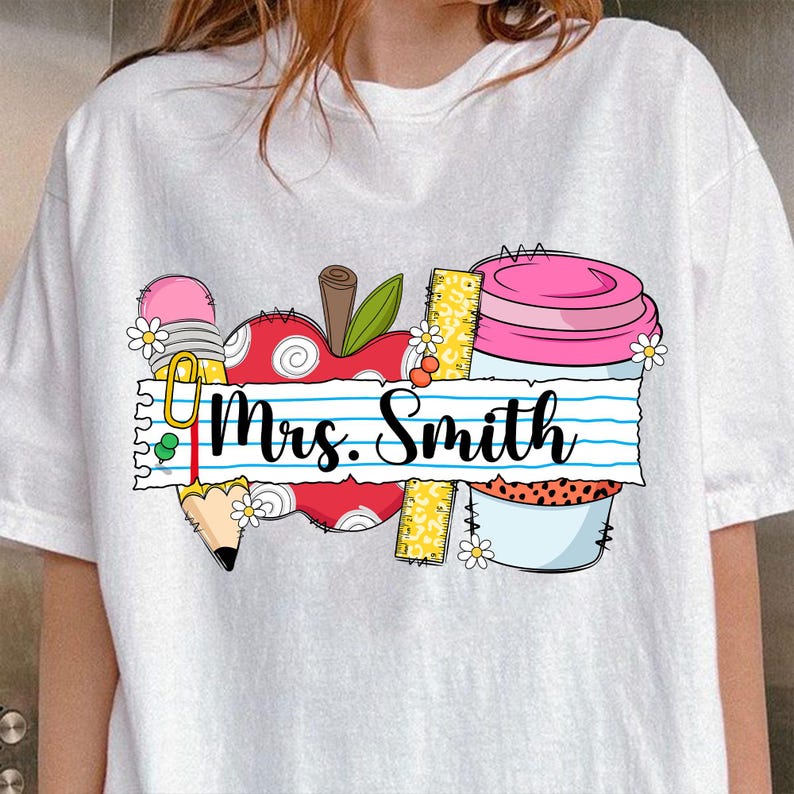 K&ouml;nnte beinhalten: Wei&szlig;es T-Shirt mit einem farbenfrohen Grafikdesign mit Elementen zum Thema Schule. Das Design umfasst einen roten Apfel, einen Bleistift, ein Lineal und einen rosa Cupcake. Der Text "Mrs. Smith" steht auf einem linierten Papierbanner.