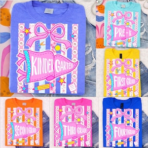 May include: A collection of colorful t-shirts featuring school grade level designs. Each shirt displays a grade level banner, a pink bow, stars, and school-themed graphics. The shirts are in various colors, including blue, orange, pink, yellow, and turquoise.
