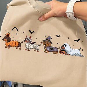 Könnte beinhalten: Beiges Sweatshirt mit einem Halloween-Motiv. Die Grafik zeigt Dackel in Kostümen: Kürbis, Mumie, Teufel, Hexe und Geist. Schwarze Fledermäuse und Sterne sind um die Hunde herum verstreut.