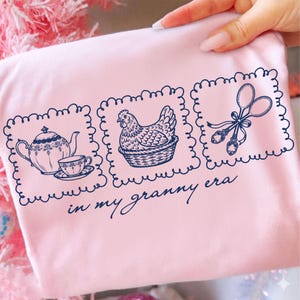 May include: A light pink t-shirt with three navy blue framed illustrations. The illustrations include a teapot and teacup, a hen in a basket, and scissors with spoons. The text "in my granny era" is below the illustrations.