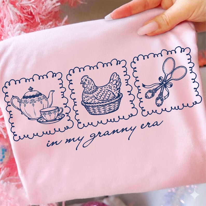 May include: A light pink t-shirt with three navy blue framed illustrations. The illustrations include a teapot and teacup, a hen in a basket, and scissors with spoons. The text "in my granny era" is below the illustrations.
