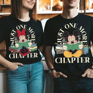 Op de afbeelding: Een zwart T-shirt met een afbeelding van Mickey Mouse en Minnie Mouse die een boek lezen. De tekst "Just One More Chapter" is in een cirkel rond de personages gedrukt.