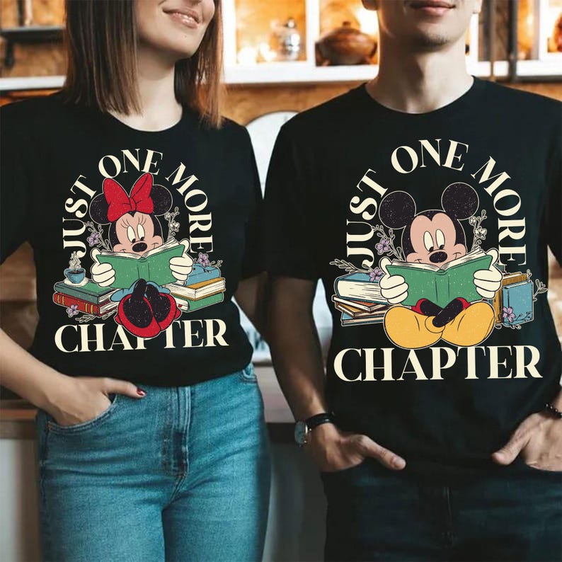 Op de afbeelding: Een zwart T-shirt met een afbeelding van Mickey Mouse en Minnie Mouse die een boek lezen. De tekst "Just One More Chapter" is in een cirkel rond de personages gedrukt.