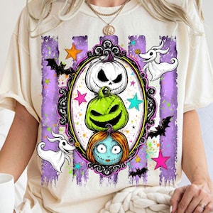 以下が含まれることがあります： クリーム色のTシャツで、装飾された黒い楕円形のフレームの中に3つのキャラクターが積み重なったグラフィックプリントが施されています。デザインには、白いカボチャ、緑のカボチャ、青い髪のキャラクターが含まれ、コウモリや星が周囲に描かれています。