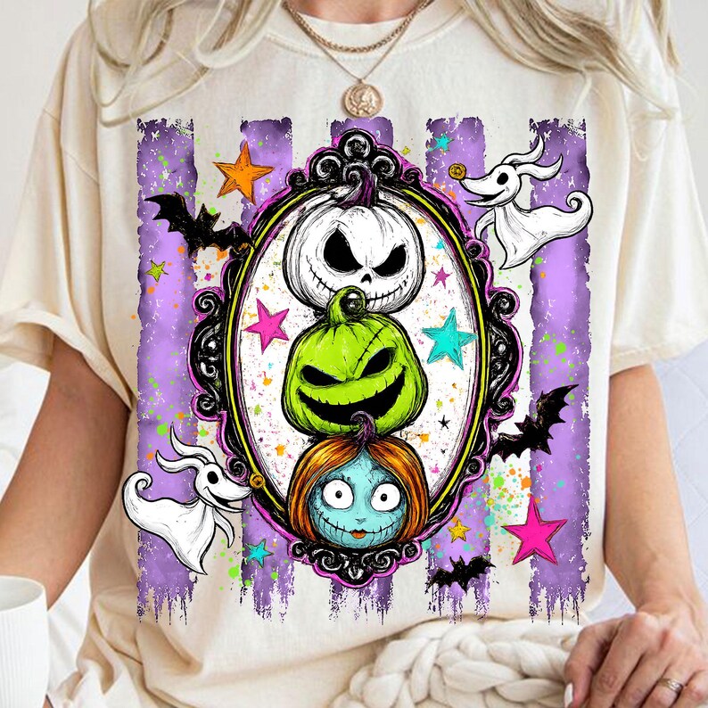 以下が含まれることがあります： クリーム色のTシャツで、装飾された黒い楕円形のフレームの中に3つのキャラクターが積み重なったグラフィックプリントが施されています。デザインには、白いカボチャ、緑のカボチャ、青い髪のキャラクターが含まれ、コウモリや星が周囲に描かれています。