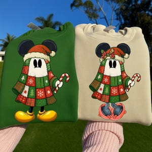 May include: Two sweatshirts, one green and one beige, featuring a cartoon character in a Christmas outfit. The character has black mouse ears, a Santa hat, and a patchwork patterned cloak. Each holds a candy cane.