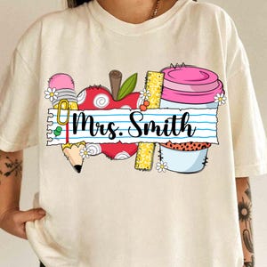 K&ouml;nnte beinhalten: Cremefarbenes T-Shirt mit farbenfrohem Grafikdesign. Das Design umfasst einen roten Apfel, einen Bleistift, ein Lineal und eine rosa Kaffeetasse. Der Text "Mrs. Smith" steht auf linierten Papier. G&auml;nsebl&uuml;mchen sind im Design verstreut.