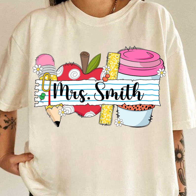 K&ouml;nnte beinhalten: Cremefarbenes T-Shirt mit farbenfrohem Grafikdesign. Das Design umfasst einen roten Apfel, einen Bleistift, ein Lineal und eine rosa Kaffeetasse. Der Text "Mrs. Smith" steht auf linierten Papier. G&auml;nsebl&uuml;mchen sind im Design verstreut.