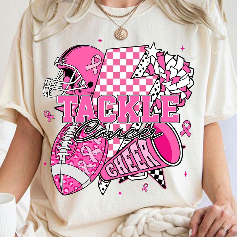 May include: Cream-coloured t-shirt with a pink and white graphic. The graphic features a football helmet, football, megaphone, and pom-pom, all in pink. The words "TACKLE Cancer CHEER" are displayed.