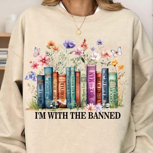 May include: A beige sweatshirt with a graphic of a bookshelf filled with banned books, including "The Bluest Eye", "Of Mice and Men", and "1984". The graphic is surrounded by colorful flowers and butterflies. The text "I'm with the banned" is printed below the graphic.