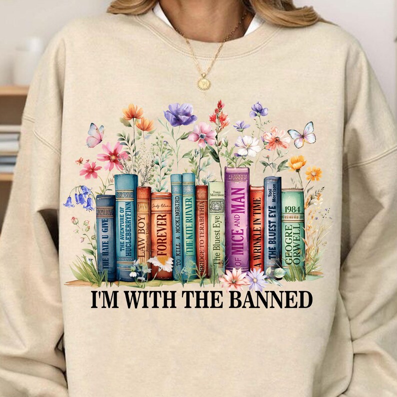 May include: A beige sweatshirt with a graphic of a bookshelf filled with banned books, including "The Bluest Eye", "Of Mice and Men", and "1984". The graphic is surrounded by colorful flowers and butterflies. The text "I'm with the banned" is printed below the graphic.