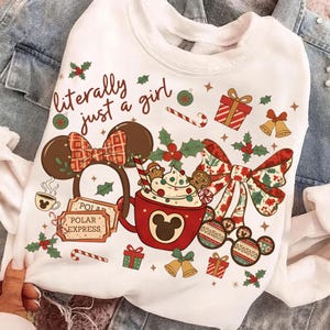 May include: White sweatshirt with a festive Christmas design. Features a red Mickey Mouse mug with whipped cream, gingerbread men, and candy canes. Includes text "literally just a girl" and holiday accents like holly and bells.