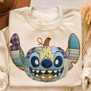 May include: Cream-colored sweatshirt featuring a patchwork design of a cartoon character's face, resembling a pumpkin. The design incorporates various blue and purple patterned fabrics, with a brown stem and a smiling mouth.