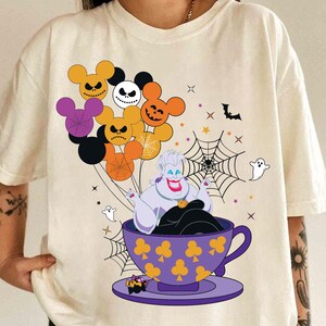 Pu&ograve; includere: T-shirt color crema con grafica a tema Halloween. Il design include una tazza viola con Ursula, che tiene in mano palloncini a forma di Mickey Mouse con Jack Skellington e altri volti spettrali. Ragnatele, pipistrelli e fantasmi completano il design.