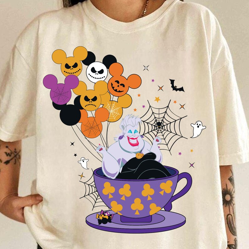 Pu&ograve; includere: T-shirt color crema con grafica a tema Halloween. Il design include una tazza viola con Ursula, che tiene in mano palloncini a forma di Mickey Mouse con Jack Skellington e altri volti spettrali. Ragnatele, pipistrelli e fantasmi completano il design.