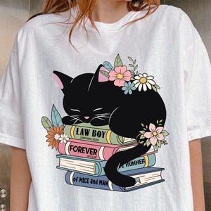 May include: A white t-shirt with a black cat sleeping on a stack of books. The books have the titles "Law Boy", "Forever", "The Runner", and "Of Mice and Men". The cat is surrounded by colorful flowers.