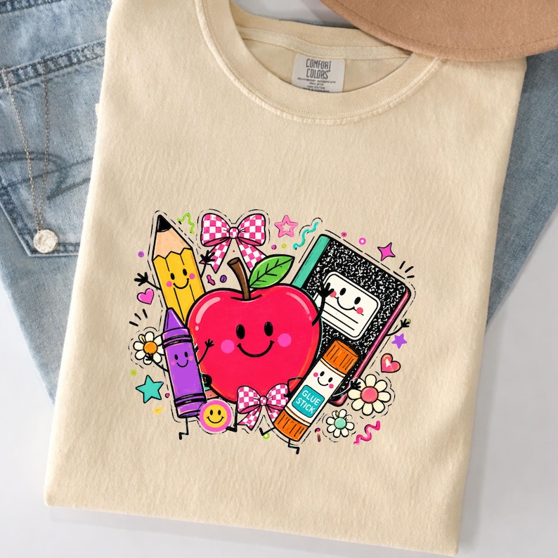 May include: A cream-colored t-shirt featuring a graphic print of a smiling red apple surrounded by school supplies including a yellow pencil, a purple crayon, a glue stick, and a black composition notebook, accented with pink checkered bows.