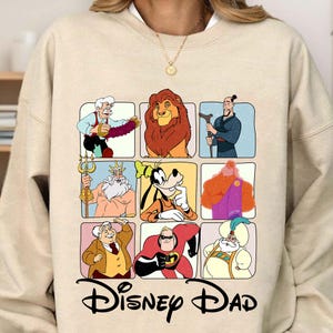May include: A beige sweatshirt with a graphic print of various Disney characters in squares. The characters include Mickey Mouse, Donald Duck, Goofy, Simba, Aladdin, Hercules, and more. The text "Disney Dad" is printed below the graphic.