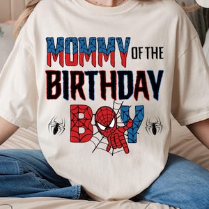 May include: White t-shirt with the text "Mommy of the Birthday Boy" in black and red. A red and blue Spiderman graphic is in the center of the shirt with two black spiders on either side.
