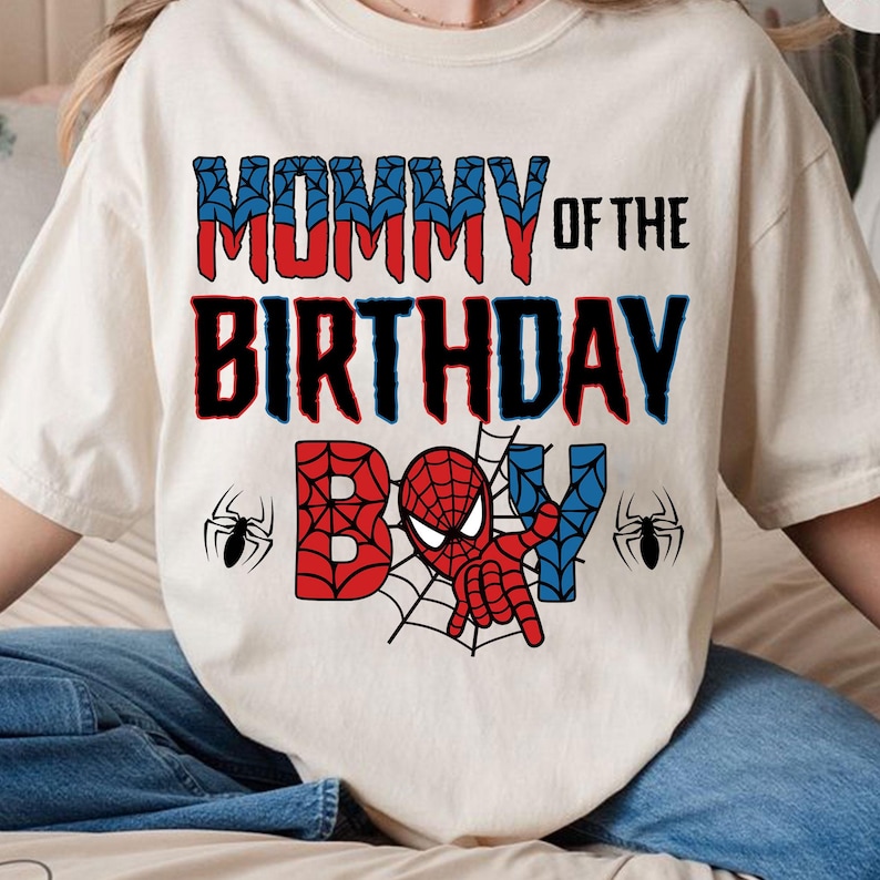 May include: White t-shirt with the text "Mommy of the Birthday Boy" in black and red. A red and blue Spiderman graphic is in the center of the shirt with two black spiders on either side.